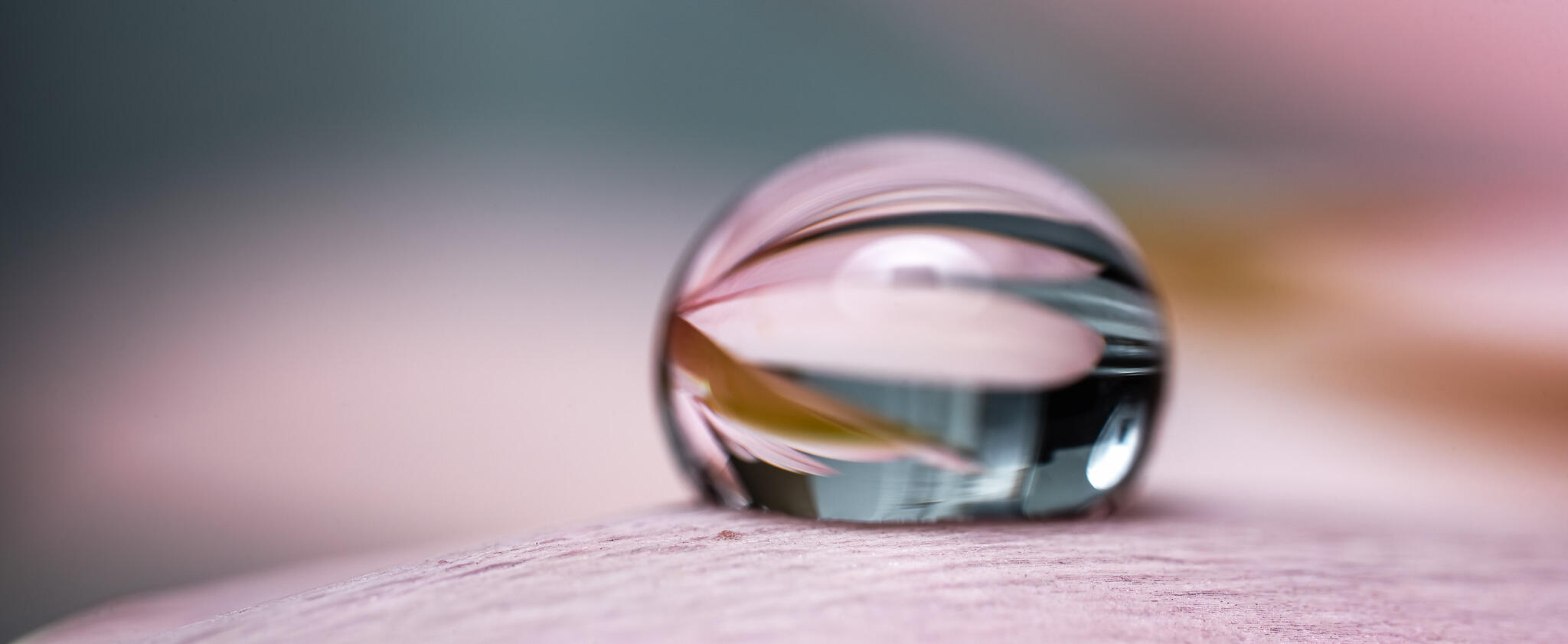 Close-up of a transparent water sphere reflecting soft pink and green tones on a neutral surface.